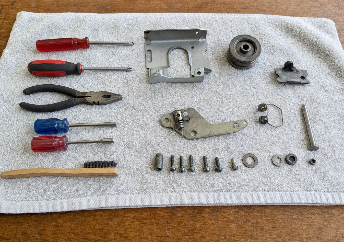 Screwdrivers, pliers, a wire brush and a wooden scraper laid on a white towel with an oven-door hinge plate, a round motor housing and loose screws